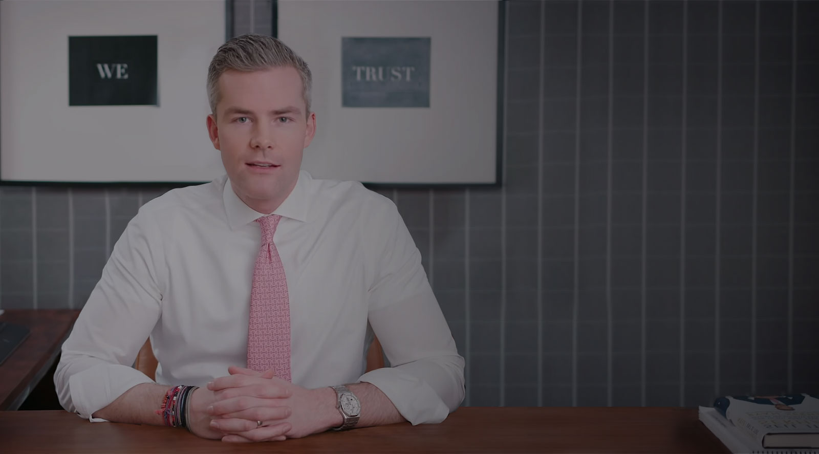 Ryan Serhant sitting at a wooden desk with his arms folded on the table.